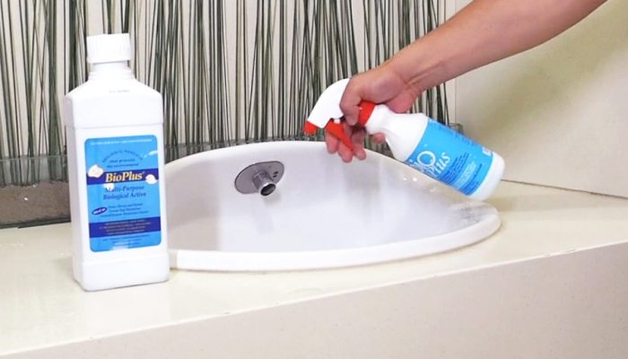 Person spraying cleaning solution on a white bathroom sink, with a disinfectant bottle nearby on the counter.