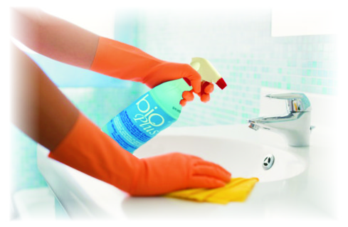 Person wearing orange gloves sprays cleaner onto a sink and wipes it with a yellow cloth in a bright bathroom.